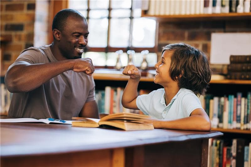 Picture of a man and child sitting at a table in a library with an open book. The man and child are fist bumping.