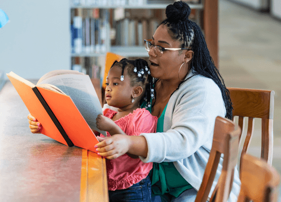 Picture of mother with young child on her lap, seated at a table in a library, reading a book together.